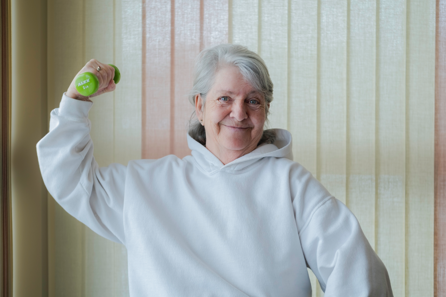 Smiling senior woman in a white hoodie lifting a green dumbbell.