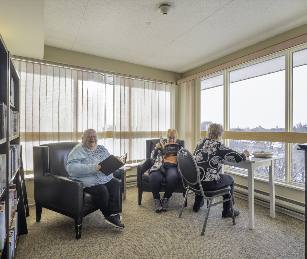 Three senior women relax in a community room, reading books and magazines, with a view of the outdoors.